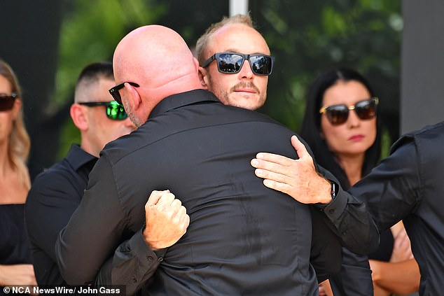 Two men hug each other dressed in black as they gather to pay their respects to the 'top gun' pilot