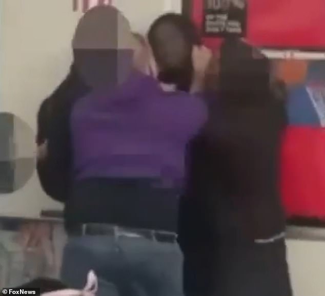 The teacher holds the student against the classroom wall while other students try to separate them.