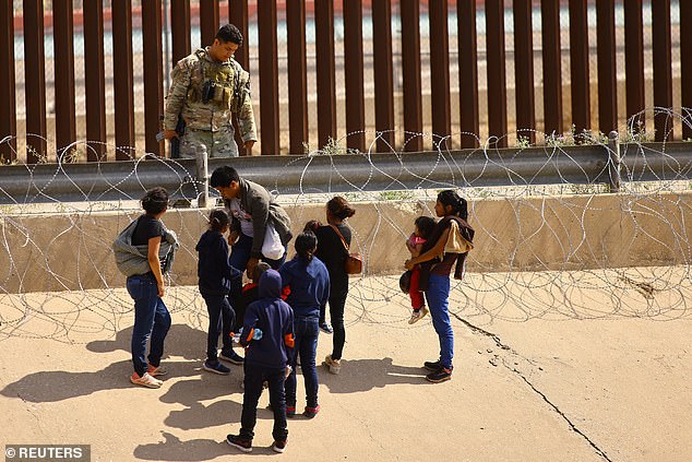 A family of migrants, with young children being held by adults, stand next to a barbed wire fence as a member of the Texas National Guard stands guard on the banks of the Rio Bravo River on August 28, 2023