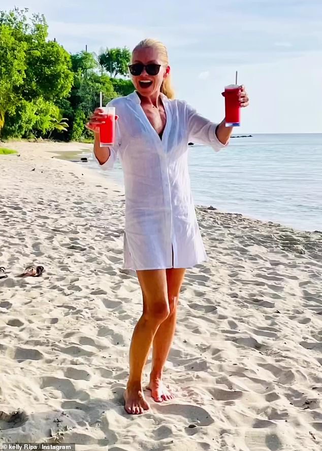 Having fun: Among the many photos, there was also a fun shot of Kelly double-fisting two colorful drinks and smiling dizzily.  She was seen barefoot in the sand, wearing a long white button-down to cover a swimsuit and large sunglasses to shield her eyes from the sun.