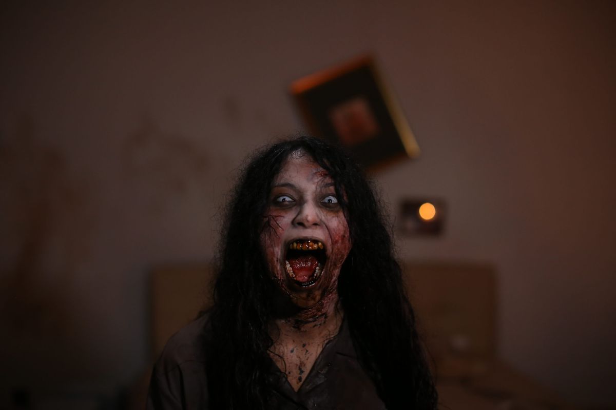 A woman with long dark hair, rotting teeth, and white skin with deep red veins, screaming in a messy room in Blood Flower.