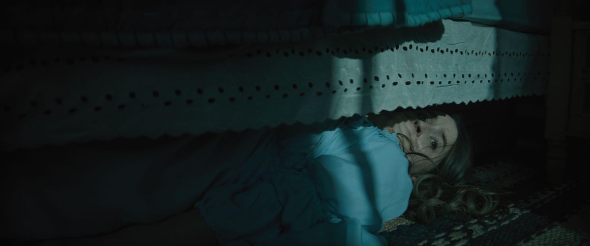 Kaitlyn Dever as Brynn hiding under a bed and covering her mouth in No One Will Save You.