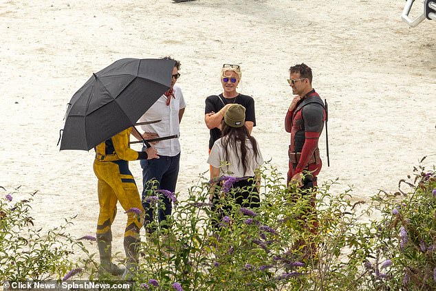 On set: The Australian actor, 54, was dressed in his yellow superhero suit as he masked his face with a large black umbrella as he chatted with his wife