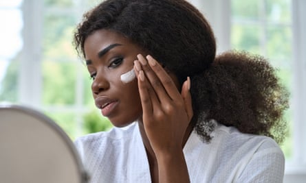 Black young woman applying cream to facial skin and looking in the mirror.