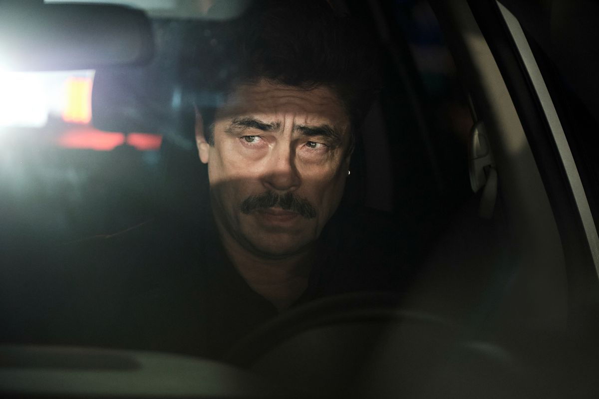 Benicio Del Toro as Tom Nichols sitting behind the wheel of a vehicle at night with headlights shining through his rear window in Reptile.