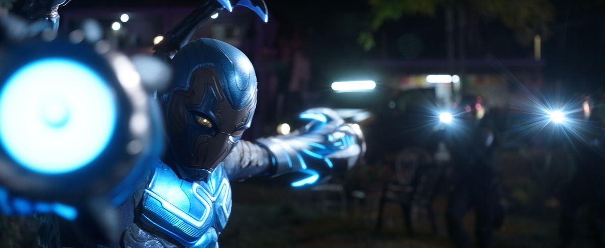 Blue Beetle poses with his arms shaped like laser cannons pointed directly toward and away from the camera in the film Blue Beetle.