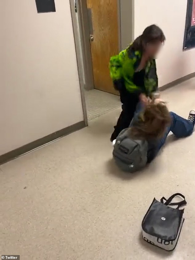 The student, wearing a green hoodie and black pants, violently pulls a smaller girl to the ground by her backpack