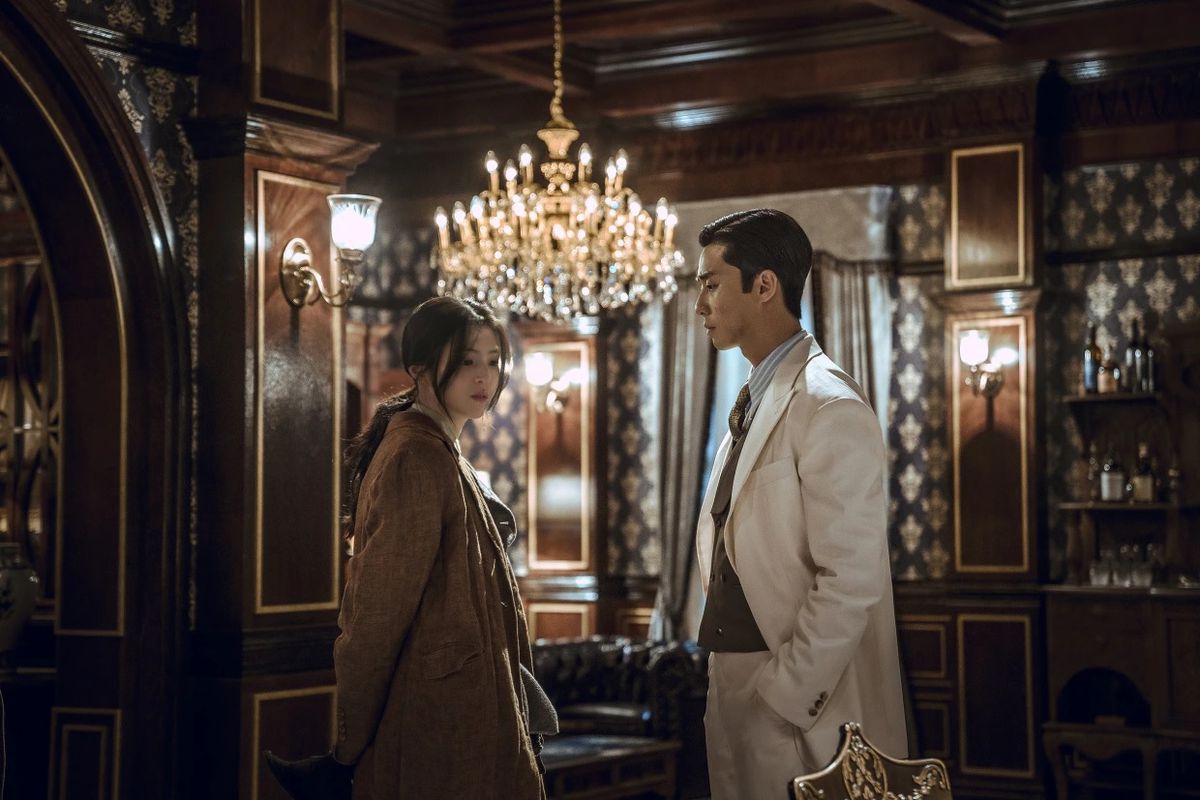 Park Seo-joon and Han So-hee wear fine clothes and stand side by side in an ornate room with a large chandelier in Gyeongsang Creature.