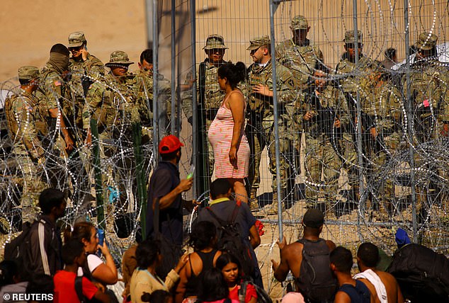 A pregnant migrant stands in front of a security fence as she is surrounded by National Guard members who have blocked the US-Mexico border crossing