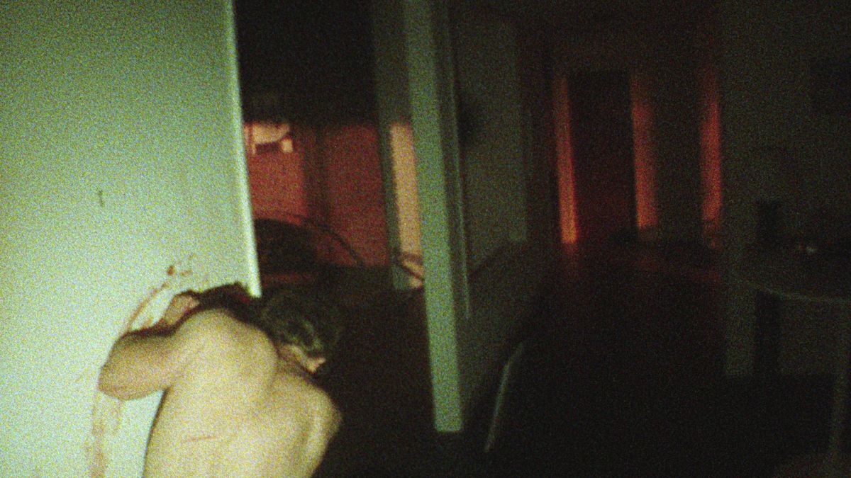 Shirtless man, seen from behind, hunched over in a dark hallway, shot granily into a dark apartment, leaning against a dirty green wall and dripping blood over it in the V/H/S/85 Dreamkill segment.