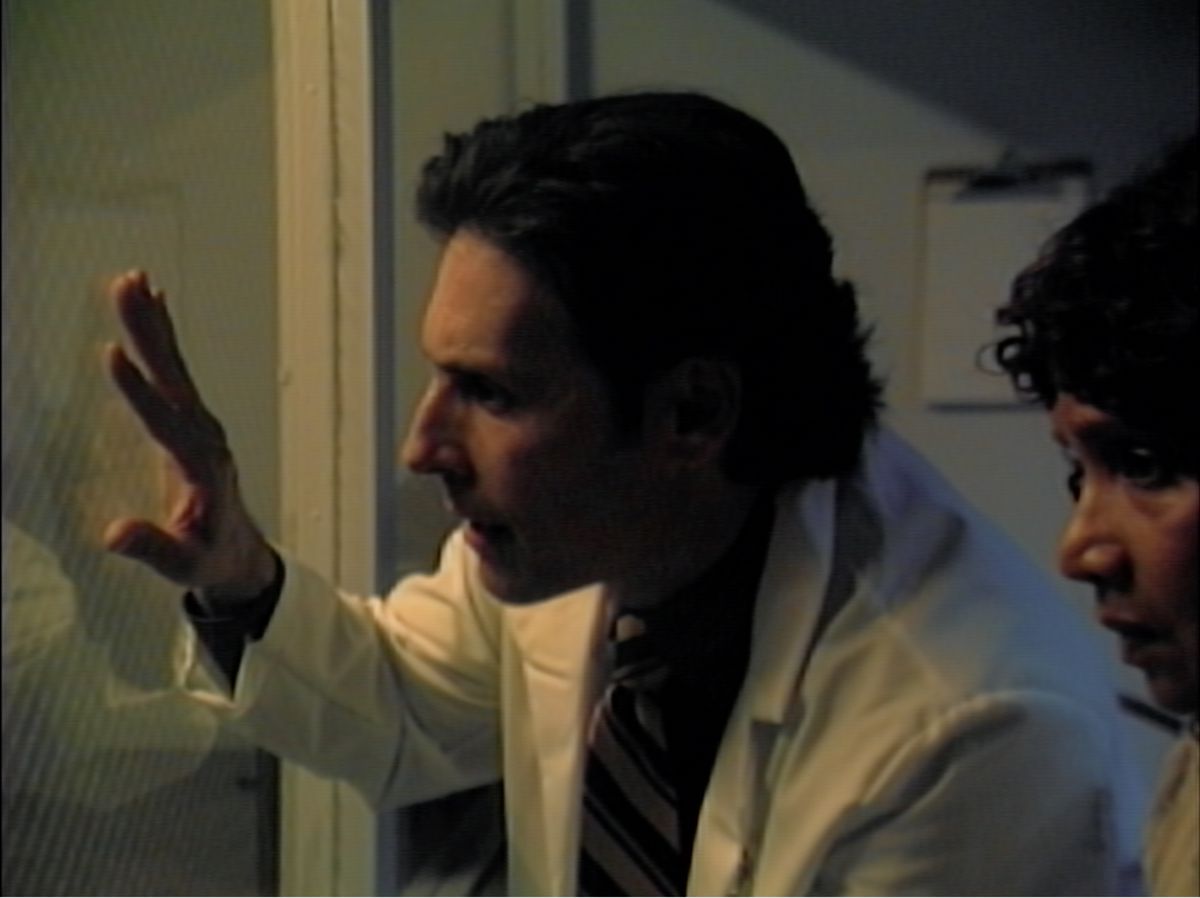 A man in a white lab coat presses his hand against the observation window and leans out as he looks into the next room in V/H/S/85 segment Total Model