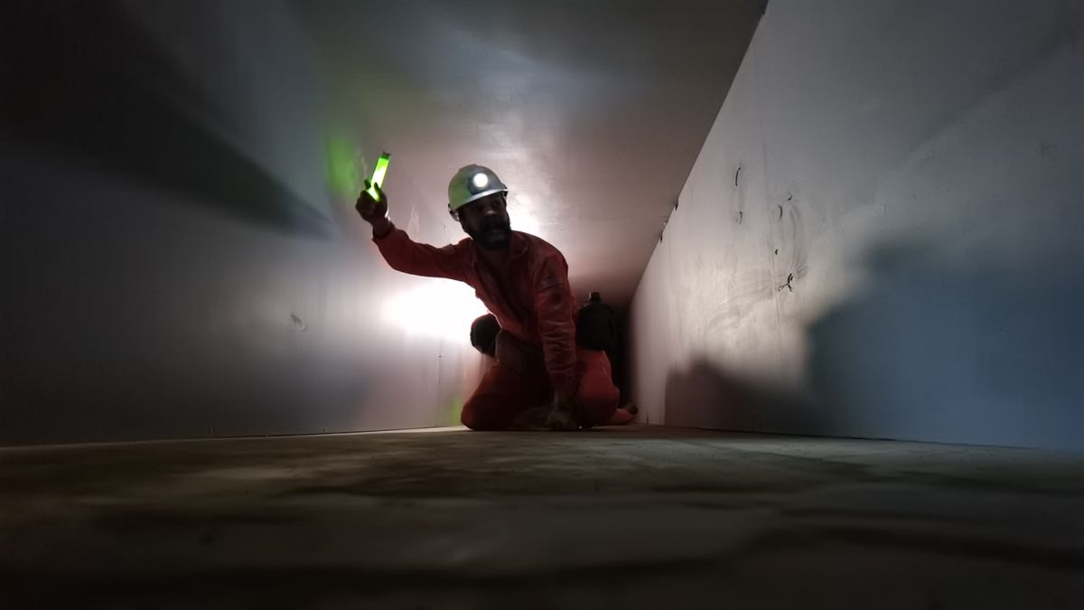 A man in an orange construction worker outfit and a yellow-green helmet with a lamp on it stands as he crawls through a white concrete tunnel in a very grainy scene from segment V/H/S/85 