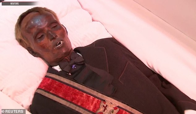 The thin man wears a black suit and tie as he lies inside a coffin at the funeral home