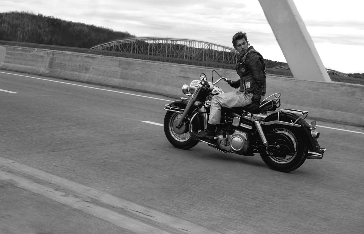 Austin Butler, in black and white, rides his Harley-Davidson over a bridge with one hand and looks back over his shoulder in a very cool way in The Bikeriders