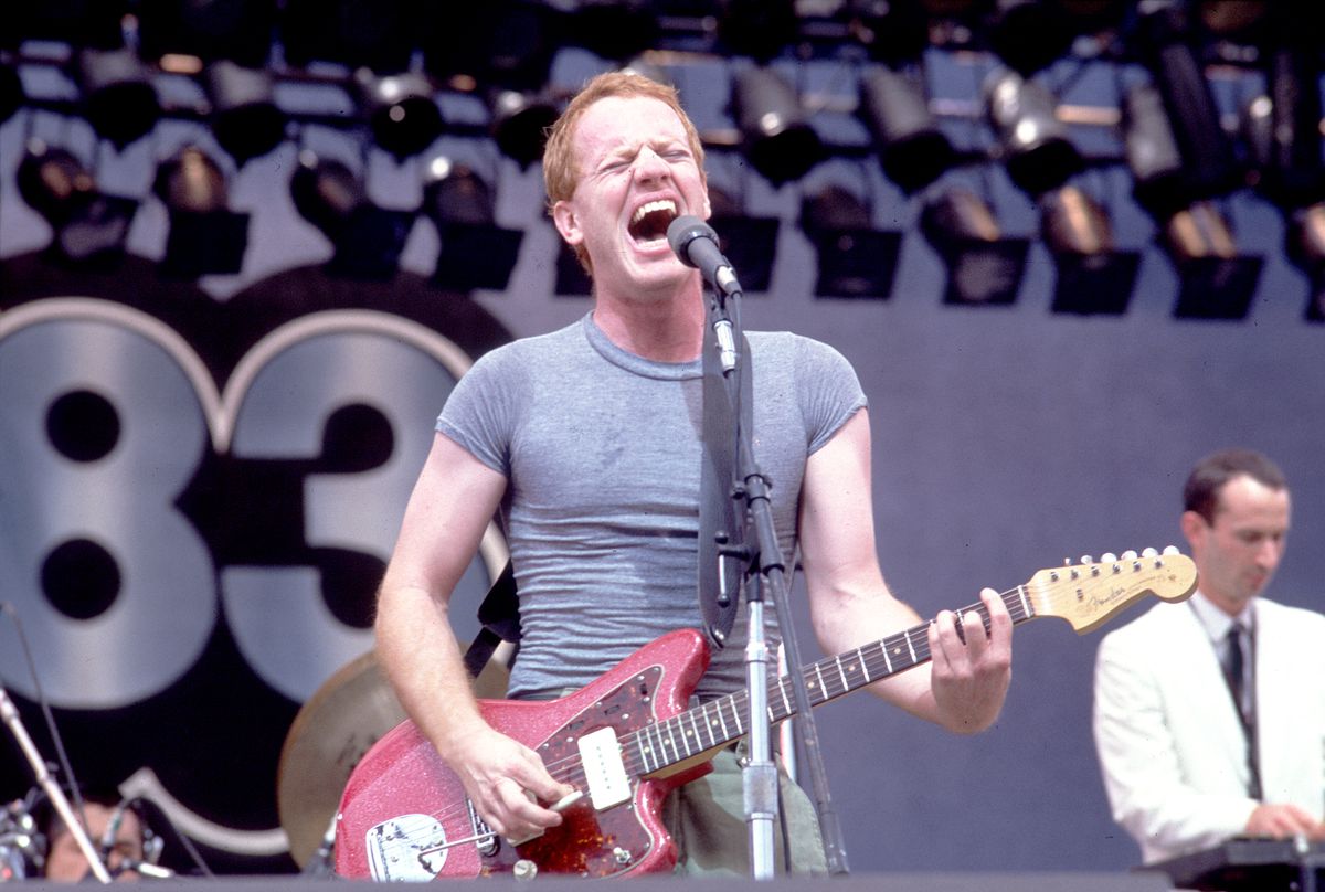 Danny Elfman, wearing a gray trunk with his hair cut short, plays an electric guitar and plays a microphone while on stage at the US Festival in Ontario, California, on May 28, 1983.