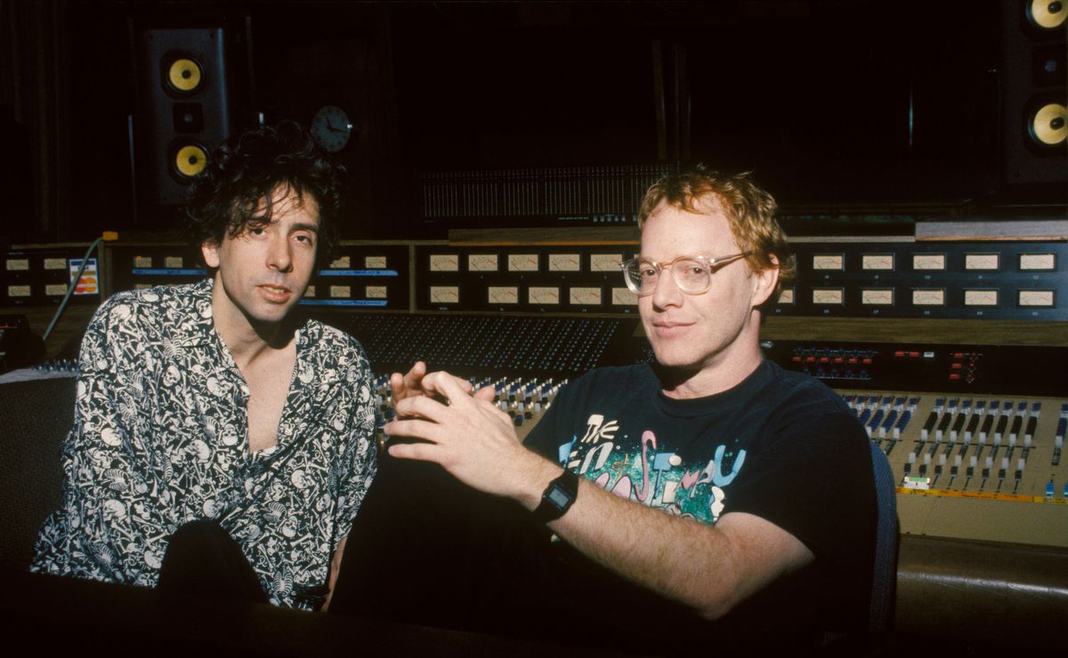 Tim Burton in a black and white button down shirt and Danny Elfman in a t-shirt, big glasses, and a dorky digital watch, sitting in the recording studio for The Nightmare Before Christmas