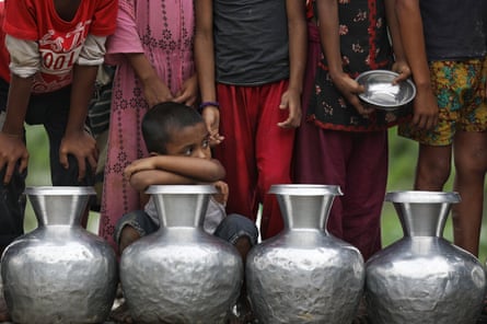 A row of children stands behind a row of metal water barrels