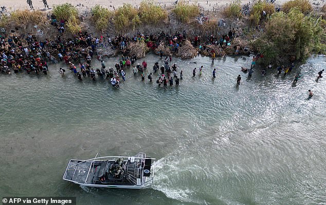 The U.S. Border Patrol boat passes a large group of migrants waiting to cross the barbed wire barrier into the United States, in Eagle Pass, Texas