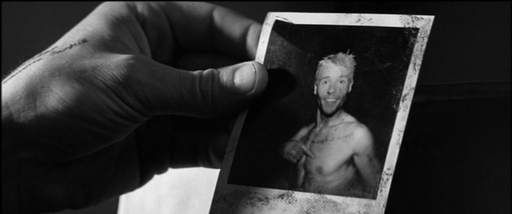 A close-up shot of a hand holding a Polaroid photograph of a man (Guy Pearce) with tattoos on his chest pointing to his heart and smiling at Memento.