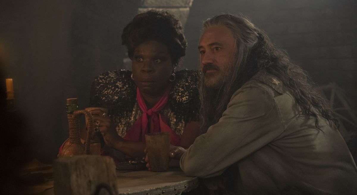 Spanish Jackie (Leslie Jones, looking horrified) and Blackbeard (Taika Waititi, looking calm) sit at a table together in Jackie's bar and watch something offscreen in season 2, part 7 of Our Death Banner.