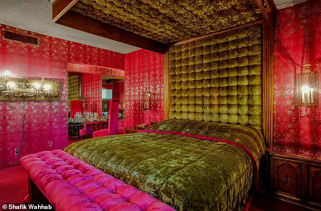 A bedroom in the house has pink carpeting and wallpaper with gold details.  There are several lamps and a dark green velvet headboard