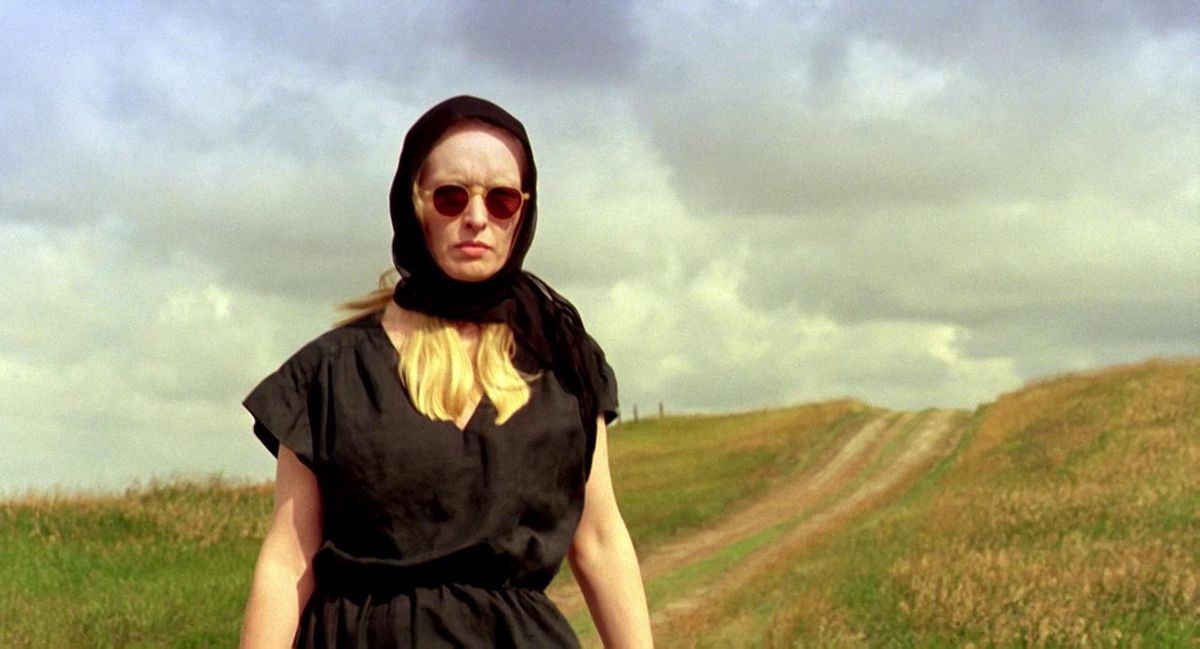 A blonde woman in all black walks down a country road in The Reflecting Skin.