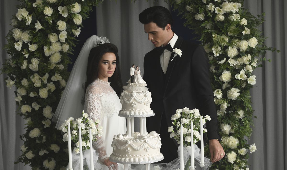 Priscilla Presley (Cailee Spaeny, in a white wedding dress and pulled back white veil), stands with Elvis Presley (Jacob Elordi, in a black tuxedo) behind their tiered white wedding cake under an arch of green leaves and white flowers, with Elvis looking at downstairs and Priscilla looks straight into the camera, in Sofia Coppola's Priscilla
