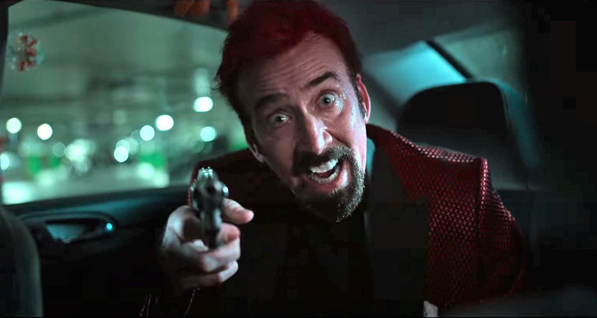 A bearded Nicholas Cage in a red suit pointing a gun from the backseat of a car and smiling maniacally in Sympathy for the Devil.