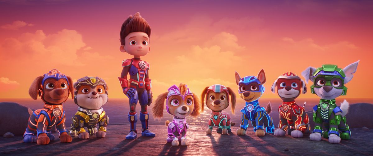 A boy in an armored suit, flanked by several dogs in similar armored suits, standing on a cliff overlooking an ocean and a sunset in Paw Patrol: The Mighty Movie.