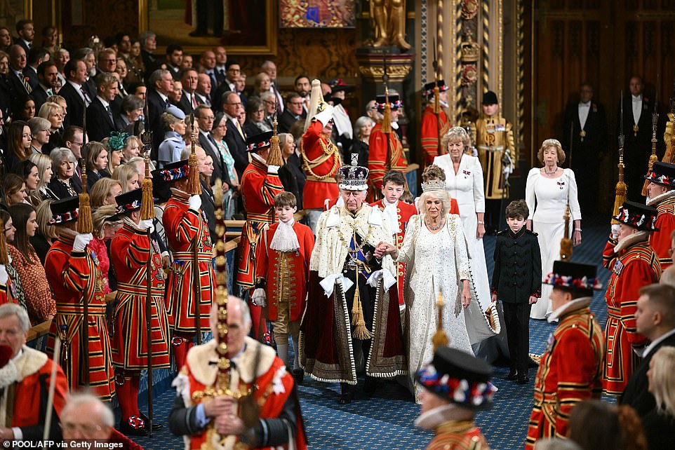 King Charles III, wearing the Imperial State Crown and State Mantle, and Britain's Queen Camilla, wearing the George IV State Diadem, walk through the Royal Gallery