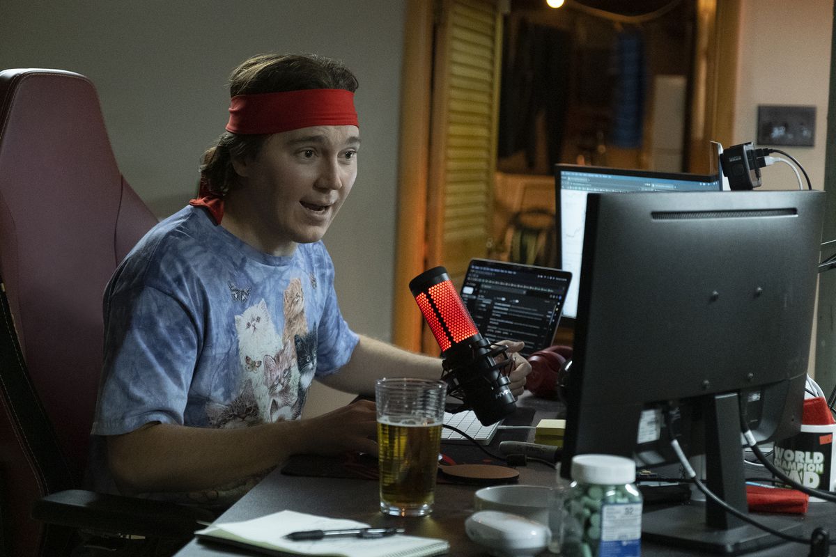 Paul Dano as Keith Gill, aka Roaring Kitty, sitting at a row of monitors at his desk with a glowing microphone in front of him, wearing a new T-shirt with fluffy kittens and a red bandana tied around his head in Dumb Money