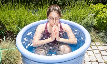 A young white woman sits up to her neck in water with ice cubes floating in it, in a blue rubber pool about waist level, on a stone patio behind some grass.