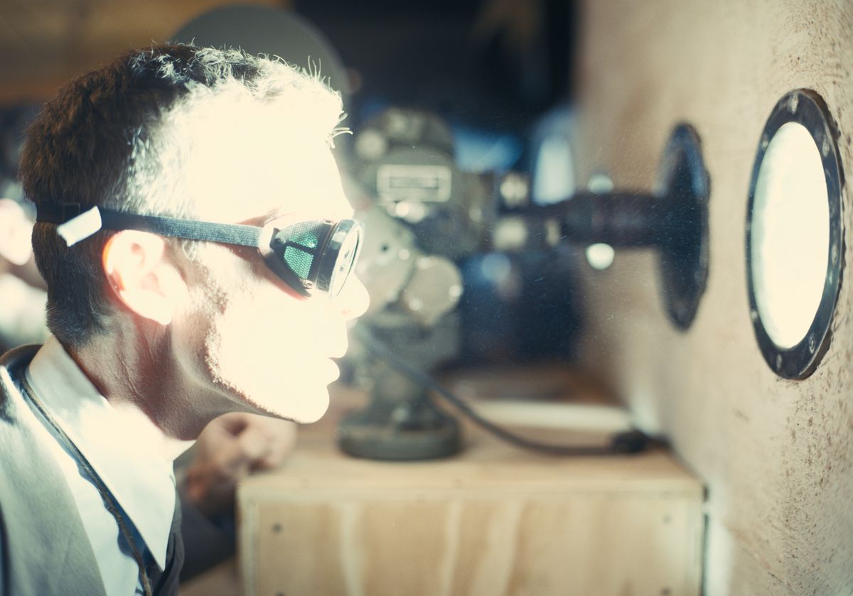 As J. Robert Oppenheimer in the film Oppenheimer, Cillian Murphy wears goggles and stares through a porthole at an explosion that casts an intensely bright light on his face