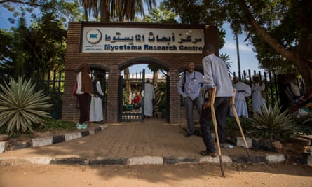 A man on crutches with one leg enters the gate of a clinic while others stand in the shade of trees