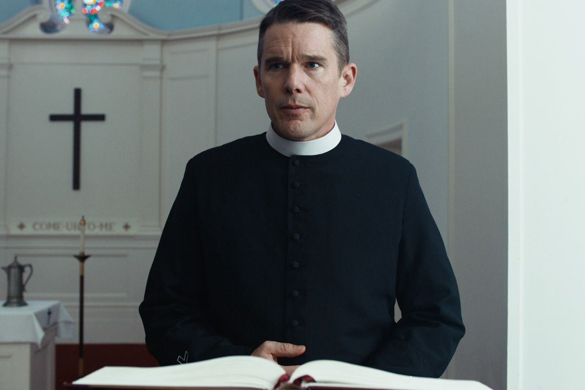 A man in priestly robes (Ethan Hawke) stands behind a lectern with a black cross visible in the background.