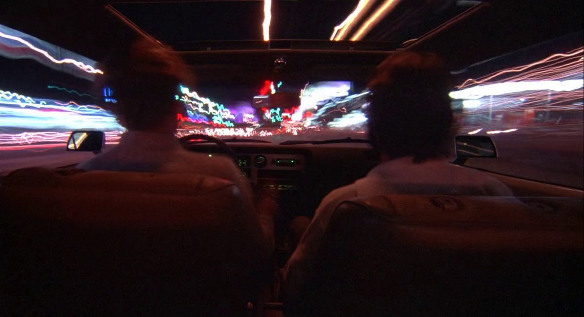 A man and a passenger behind the wheel of a car as streaks of light shoot around them in Koyaanisqatsi.