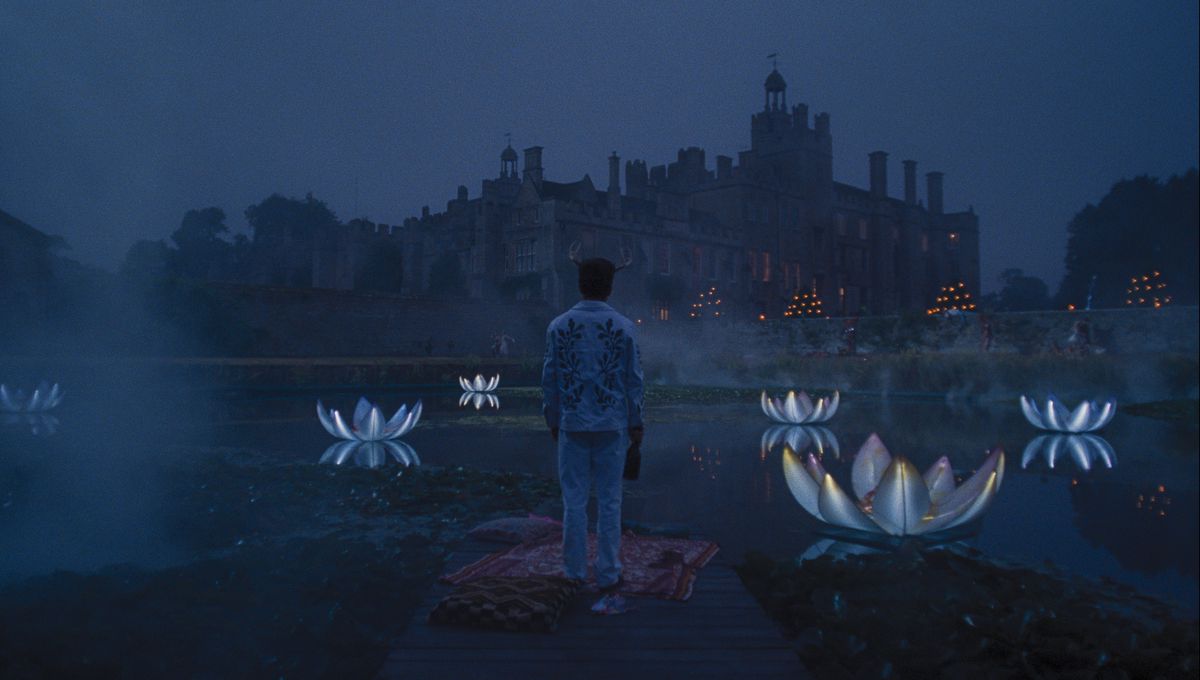 Oliver (Barry Keoghan), dressed for a costume party in deer horns and a white suit with lots of beads, stands on a blanket on a dock with his back to the camera and looks out over a pond full of enormous glowing floating plastic lilies.  and beyond, an immense Gothic estate, in the film Saltburn