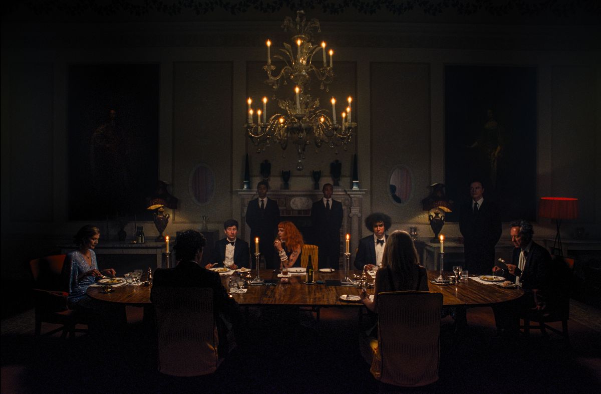 Oliver (Barry Keoghan) sits at a long dining table in a very dark room with Felix (Jacob Jacob Elordi) and other members of his family, all in formal clothes, in the film Saltburn