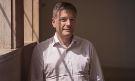 Brian Bell, in a cotton shirt that is slightly wrinkled towards the waist, stands smiling by a sunny window