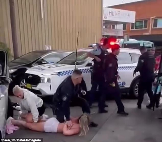 The woman, dressed in a tank top and shorts, can be heard shouting at a man who is also being held back by other officers