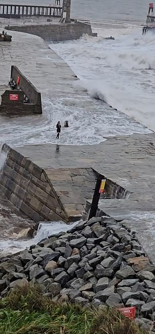 This is the moment an 'idiot' tourist taking a selfie on top of a stone pier was caught off guard by a colossal wave that washed over the wall of the East Pier in Whitby, North Yorkshire, on Tuesday.