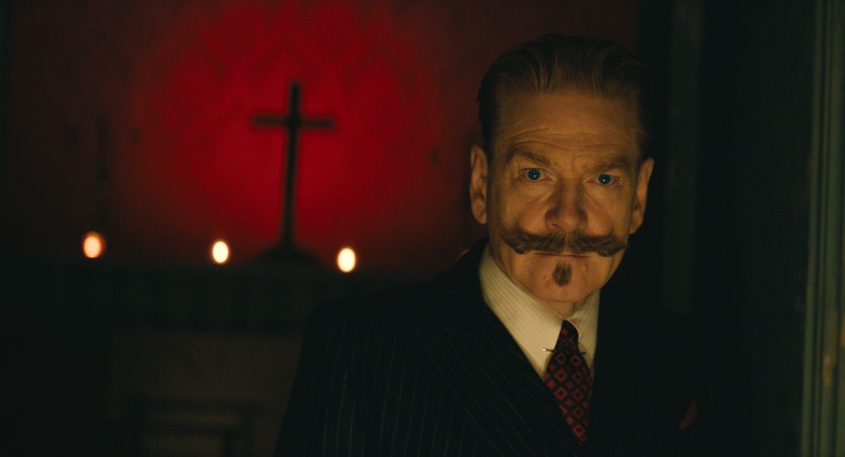 Hercule Poirot by Kenneth Branagh, standing in a dark room.  A cross hangs on the wall behind him.