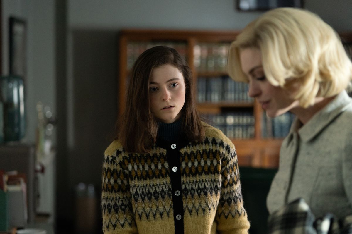 Thomasin McKenzie stands in some kind of ugly brown sweater and nervously watches the blonde Anne Hathaway doing glamorous office work in the film Eileen