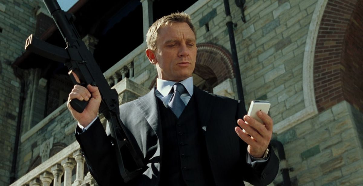 Daniel Craig as James Bond in Casino Royale, wearing a suit and looking at his phone while holding a gun