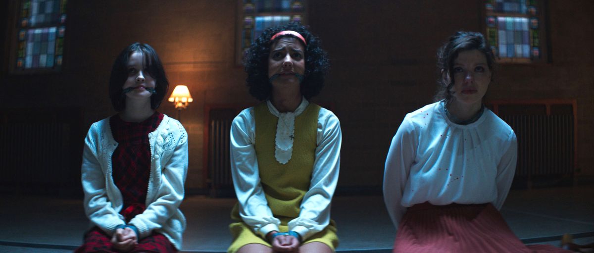 Clara (Georgia Acken) and Samantha (Madison Baines) kneel with their hands tied in front of them and gagging in their mouths, next to their teacher Rose (Chloë Levine), kneeling with her hands tied behind her back in The Sacrifice Game