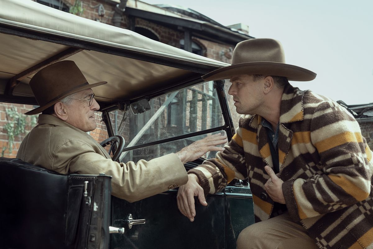 Leonardo DiCaprio in a cowboy hat and poncho leans over a car driven by Robert DeNiro in Killers of the Flower Moon.