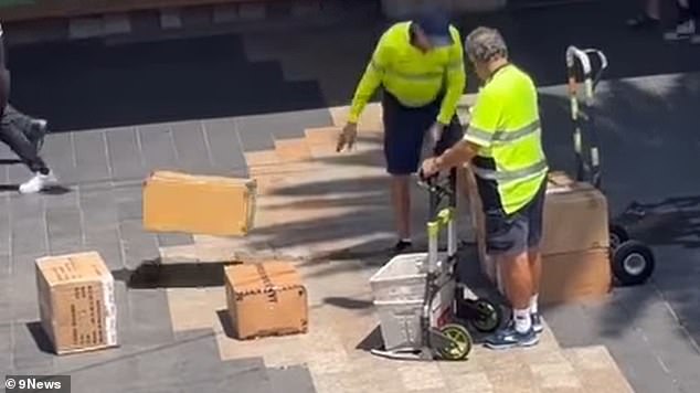 The frustrated postman grabbed boxes from the cart and threw them while the older postal worker still watched