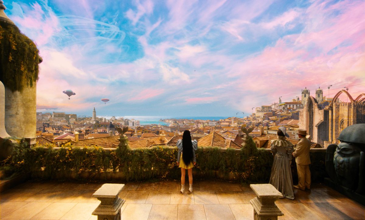 Emma Stone as Bella Baxter stands in front of a fantastic, colorful view of the city of Lisbon in Poor Things