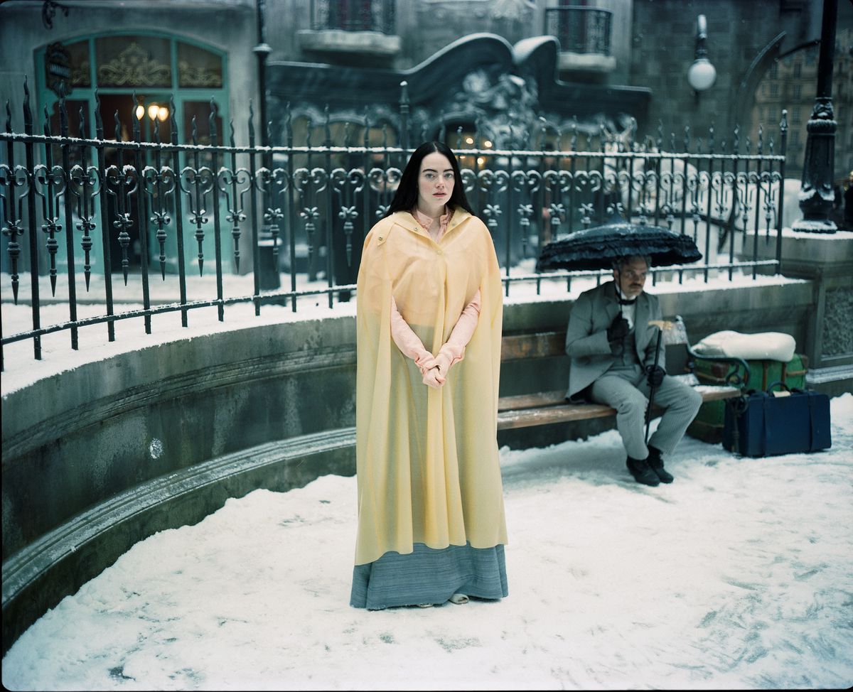 Emma Stone as Bella Baxter stands in the snow, with a depressed looking Mark Ruffalo under an umbrella behind her in Poor Thigns