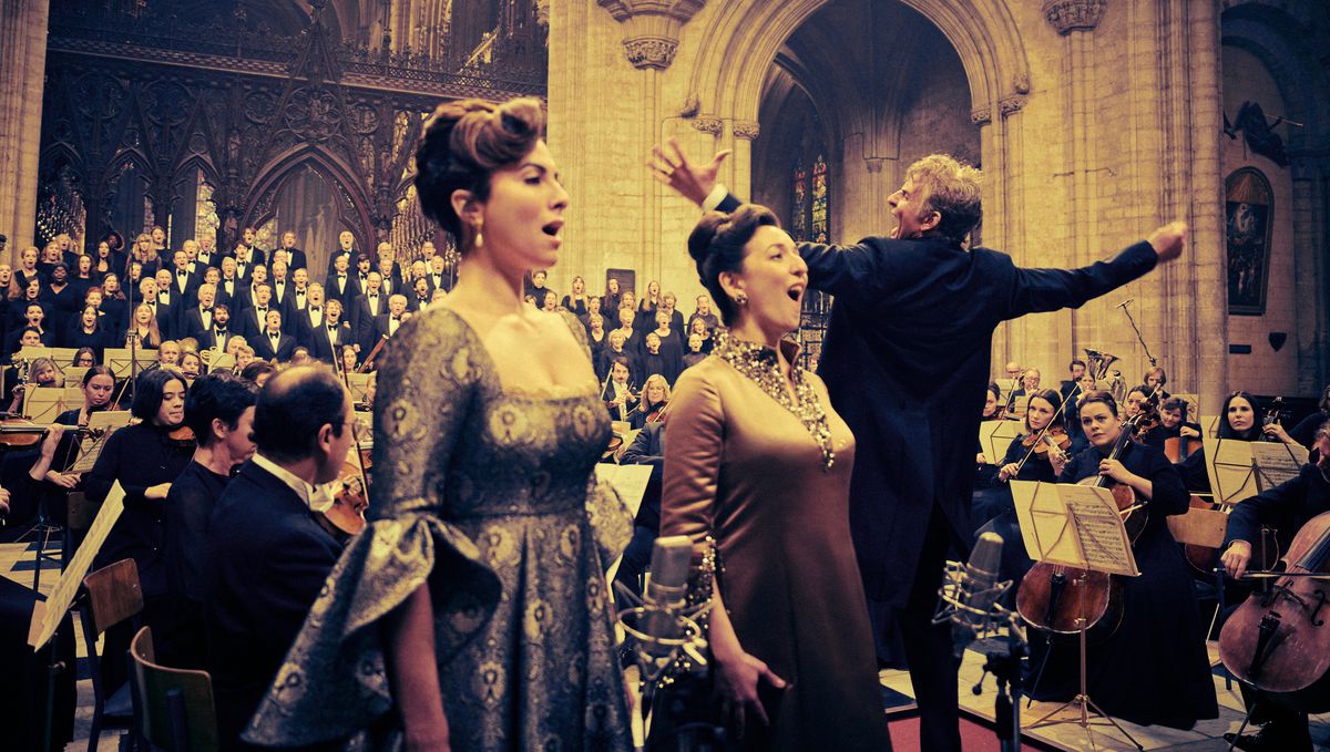 Bradley Cooper as Leonard Bernstein conducts flamboyantly in a cathedral as two operatically dressed soloists (Isabel Leonard and Rosa Feola) sing to him, an orchestra flanks him and a choir of men in tuxedos sing in the background in Netflix's Maestro